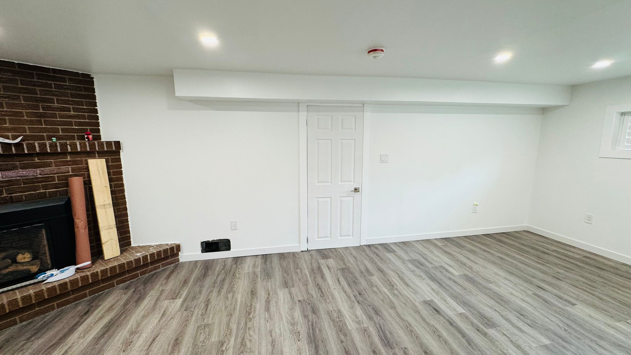 Finished basement family room with white walls, oak flooring and a brick fireplace